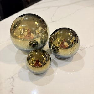 Op de afbeelding: Drie bolvormige objecten met een sterk reflecterende, metallic gouden afwerking. De bollen variëren in grootte, met één grote, één middelgrote en één kleine bol. Elk glad, gepolijst oppervlak fungeert als een spiegel en reflecteert de omgeving.