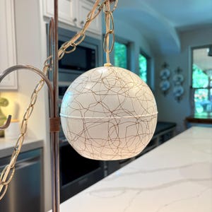 May include: A spherical pendant lamp featuring a white shade with a brown, thin, chaotic line pattern. The lamp is suspended by a metallic chain attached to a vertical metal stand. The design consists of two hemispherical halves joined together.