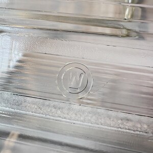 May include: Close-up of a clear glass object with a ridged surface. A circular emblem with the letter "W" is embossed on the glass. The object has a reflective quality, with light reflecting off the ridges and the emblem.