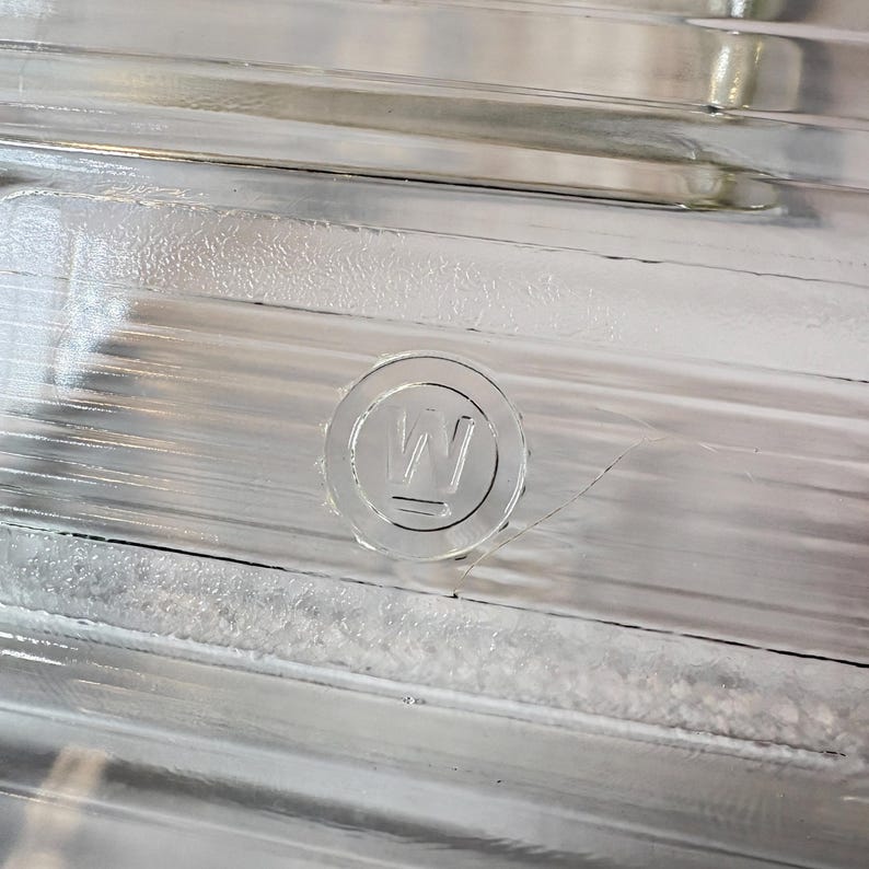 May include: Close-up of a clear glass object with a ridged surface. A circular emblem with the letter "W" is embossed on the glass. The object has a reflective quality, with light reflecting off the ridges and the emblem.