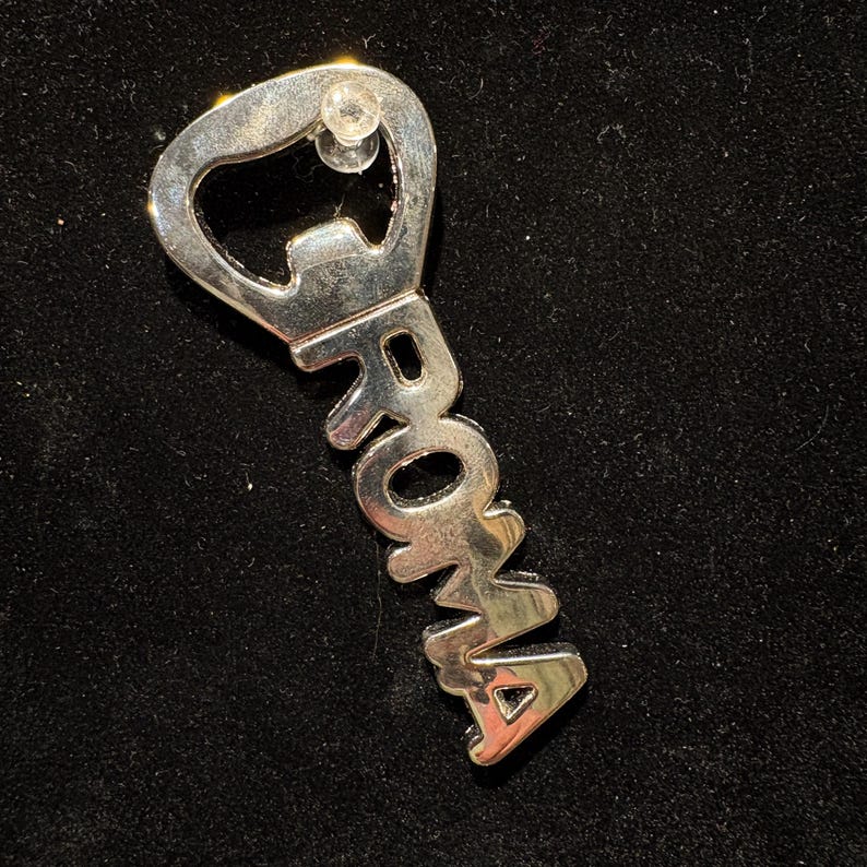 May include: A silver bottle opener with the word "ROMA" in capital letters. The opener has a key-like shape, with a bottle cap opener at the top. The word "ROMA" is in a stylized font.