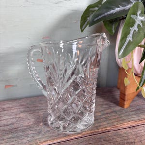 May include: A clear glass pitcher with a diamond pattern. The pitcher has a handle and a spout.