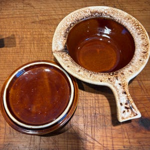 May include: A brown and white glazed ceramic casserole dish with a lid. The dish has a handle and a brown interior.