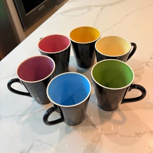 May include: A set of five black ceramic mugs, each featuring a distinctively colored interior. The mugs have interiors in shades of pink, yellow, light yellow, lime green, and light blue, all with a glossy finish and a matching black handle.