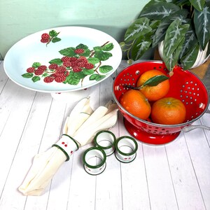 May include: A white serving platter with a raspberry design, a red colander with oranges, folded white napkins with cherry-decorated napkin rings. A decorative kitchenware set.