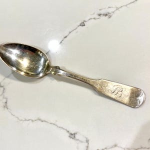 May include: A silver spoon with a rounded bowl and a long handle. The handle has a decorative design near the bowl and is engraved with the letters "PB". The spoon is likely vintage or antique.