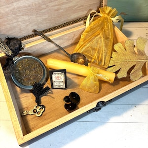 May include: A wooden box with various items, including a decorative spoon, a candle, a small bag, a coin, a key, and a small box with text. The items are arranged inside the box, creating a vintage aesthetic.