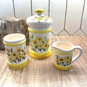 May include: A ceramic kitchen set featuring a canister with a lid, a small pitcher, and a cylindrical container. Each piece is white with yellow accents and a floral daisy design. The set is displayed on a wooden surface.