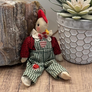 Może przedstawiać: Ręcznie robiona materiałowa lalka koguta z czerwonym grzebieniem, muszką i ogrodniczkami w kratę. Lalka ma kremową twarz i ma na sobie czerwono-zielony strój w kratę. Lalka siedzi na drewnianej powierzchni.