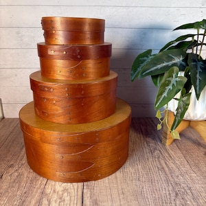 May include: A set of four round, graduated wooden boxes stacked vertically. Each box features a smooth, medium-brown finish with visible wood grain and overlapping side bands secured by small, evenly spaced metal fasteners along the curved edges.
