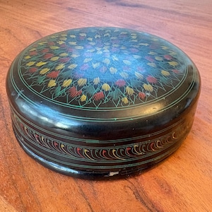 May include: A round black lacquered box with a colourful floral design. The lid is decorated with a mandala-like pattern in shades of red, yellow, and green.