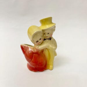 Può includere: Statuina in ceramica vintage di una coppia che si abbraccia, con la donna in un abito rosso e l'uomo in un completo giallo e cilindro. La statuina ha una piccola apertura, suggerendo che potrebbe essere un vaso o una fioriera. La coppia è in un abbraccio affettuoso.