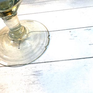 May include: A clear glass wine glass with a round base and a long stem. The base has a slightly uneven edge. The word "OTOGIE" is printed on the base. The glass is set against a white wooden surface.