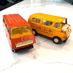May include: Two vintage toy buses. One is orange, the other is yellow with the text "TONKA SCHOOL DISTRICT 277". Both have black wheels and white tires. The orange bus has a white grill and a yellow windshield.