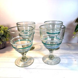 May include: Four clear glass goblets with a footed base. Each goblet has a wide bowl with horizontal stripes of teal, white, and light blue. The glasses are arranged on a light-colored surface, suggesting a set for serving beverages.