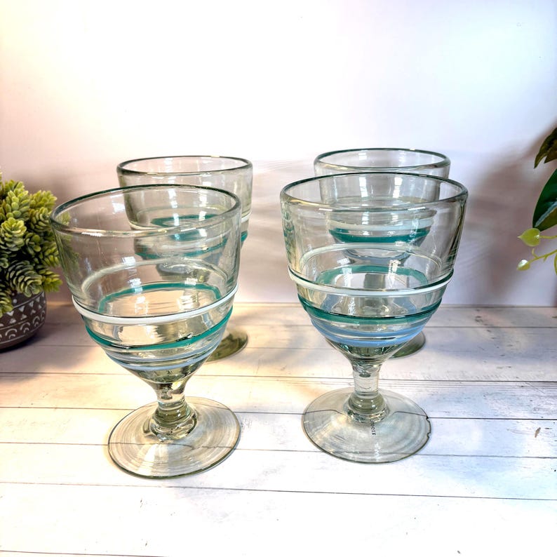 May include: Four clear glass goblets with a footed base. Each goblet has a wide bowl with horizontal stripes of teal, white, and light blue. The glasses are arranged on a light-colored surface, suggesting a set for serving beverages.
