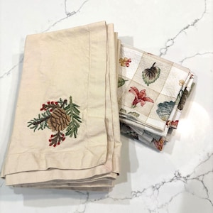 以下が含まれることがあります： ベージュ色の布ナプキン。松ぼっくりと葉の刺繍が施されています。刺繍入りのナプキンの隣には、花柄と葉柄のナプキンが重ねられています。ナプキンは折りたたまれ、白い大理石の表面に積み重ねられています。