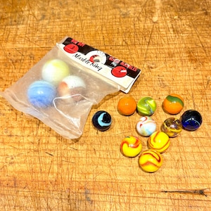 Peut inclure: Un ensemble de billes de verre colorées, certaines dans un sac en plastique transparent, dispersées sur une surface en bois. Le sac contient des billes dans les tons de jaune, bleu et blanc. D'autres billes sont orange, vertes et jaunes avec divers motifs. L'emballage indique "Marble King".