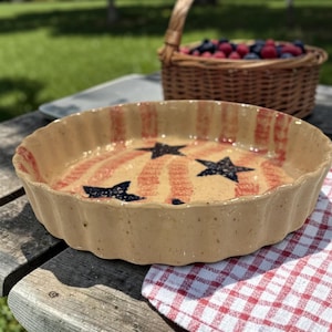 May include: A round, beige ceramic pie dish with a scalloped edge. The interior features red stripes and navy blue stars. A woven basket of berries sits in the background, suggesting a picnic setting.