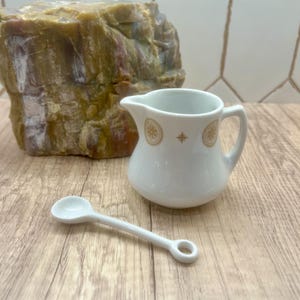 May include: A white ceramic creamer with a handle and a small white spoon. The creamer has a gold design and a spout for pouring. The spoon is small and has a round bowl. The creamer and spoon are on a wooden surface.