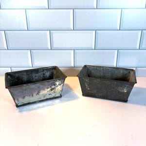 May include: Two rectangular metal planters with a distressed, aged appearance. The planters are dark gray with areas of lighter, silvery tones, suggesting wear and age. The design is simple and utilitarian.