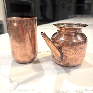 May include: Two copper vessels are displayed. One is a tall, cylindrical cup. The other is a small, round pot with a spout. Both have a shiny, metallic finish and show signs of use. The items are likely for kitchen or decorative use.