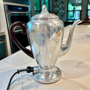 以下が含まれることがあります： ダークブラウンのハンドルと黒い電源コードが付いた、ヴィンテージのシルバー電気コーヒーポット。 ポットには透明なガラスノブと注ぎ口が付いた蓋が付いています。 この電化製品は白いカウンタートップに置かれています。