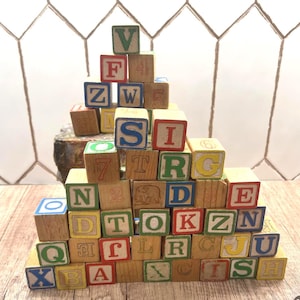 Op de afbeelding: Een verzameling houten alfabetblokken, sommige met cijfers, gestapeld in een piramidevorm. De blokken bevatten letters in verschillende kleuren, waaronder rood, blauw, groen en geel. De blokken staan op een houten oppervlak.