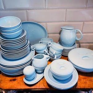 May include: A complete light blue ceramic dinnerware set. The set includes bowls, plates, cups, a serving platter, a creamer, a sugar bowl, a covered dish, and a pitcher with a lid. The dishes are stacked on a wooden surface.