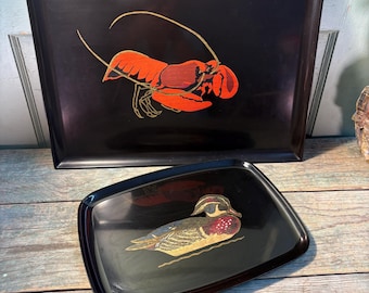 May include: Two black serving trays. The larger tray features a red and gold lobster design. The smaller tray has a duck illustration in gold, red, and blue. Both trays have a glossy finish.