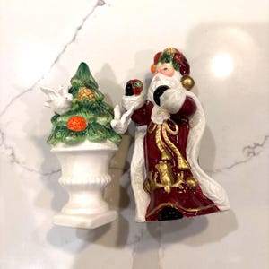 May include: Two ceramic Christmas figurines. One is a green Christmas tree with white doves and an orange ornament on a white urn. The other is a Santa Claus figure in a red coat with gold accents and a white beard.