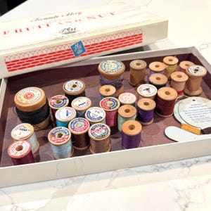 Puede incluir: Una colección de carretes de hilo de madera vintage en varios colores, incluyendo negro, azul, morado, rosa y marrón, guardados en una caja de dulces Fannie May Fruit and Nut. Los carretes tienen etiquetas de marcas como Coats and Clark.