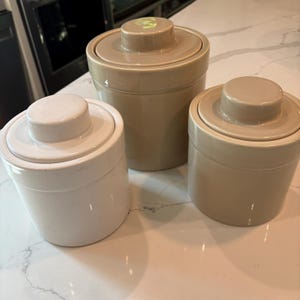 Pode incluir: Um conjunto de três potes cilíndricos de cerâmica com tampas correspondentes, cada um com um puxador circular elevado. Um pote é de um branco brilhante, enquanto os outros dois são de um tom bege neutro, dispostos juntos em um grupo.