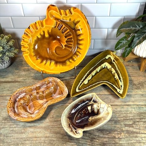 May include: Four vintage ceramic ashtrays in shades of brown, yellow, and green. The ashtrays have unique shapes, including a large, spiral-shaped one and a triangular design. The collection is displayed on a wooden surface.