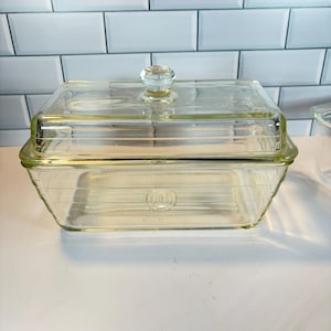May include: A clear glass rectangular butter dish with a lid and a faceted knob. The dish has a ribbed design and sits on a white surface. The background features a white tiled wall.
