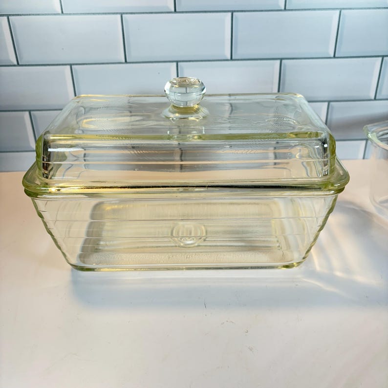 May include: A clear glass rectangular butter dish with a lid and a faceted knob. The dish has a ribbed design and sits on a white surface. The background features a white tiled wall.