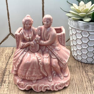 May include: A pink ceramic planter featuring a seated couple in period clothing. The man holds a lute, and the woman is seated beside him. The planter has a rectangular opening and is decorated with ornate details. A succulent plant in a white pot is in the background.