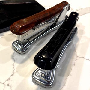 May include: Two vintage staplers. One has a brown, marbled handle, the other is black. Both have a shiny, silver base and the word "Ace" is printed on the top of each stapler.