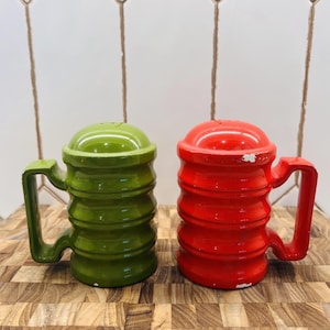 May include: A pair of vintage salt and pepper shakers. One is green, the other is red. Each shaker has a handle and a ridged body. The shakers are on a wooden surface.