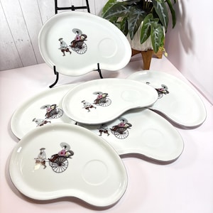 Puede incluir: Juego de seis platos de servir de cerámica blanca con borde negro y dorado. Cada plato presenta una ilustración en blanco y negro de una persona en un rickshaw con flores rosas.