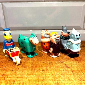 May include: A collection of vintage cartoon character figurines, including Donald Duck, a green dinosaur, Fred Flintstone, Barney Rubble, a wolf, and a robot. The figurines are made of plastic and are brightly colored.