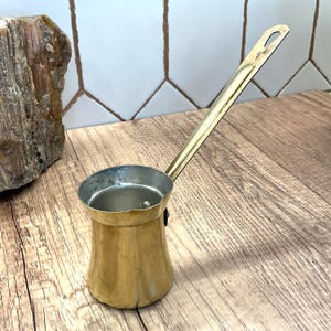 May include: A brass-colored Turkish coffee pot, also known as a cezve, with a long handle. The pot has a wide base that tapers to a narrow neck and a flared rim. The handle is attached to the side of the pot and has a loop at the end for hanging.