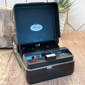 May include: A black portable Mercury cassette recorder with an open lid. The device features a red record button, a black play/rewind/advance control switch, and a silver trim. The interior lid displays the Mercury brand logo in an oval.