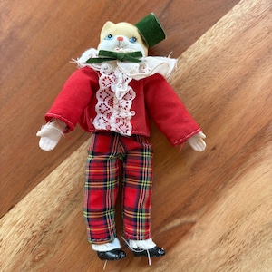 May include: A porcelain doll with a cat face wearing a red jacket with white lace trim, a green bow tie, a green hat, red and black plaid pants, and black shoes.
