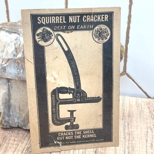 以下が含まれることがあります： Squirrel Nut Crackerのヴィンテージ広告。長方形の紙に道具の白黒イラストが描かれています。テキストには「SQUIRREL NUT CRACKER」「BEST ON EARTH」「ALEX WOLDERT CO.」「CRACKS THE SHELL BUT NOT THE KERNEL」と記されています。