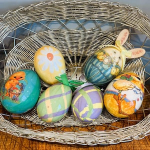 May include: Six decorated Easter eggs in a silver wire basket. The eggs are decorated with various designs, including a bunny, a chick, a flower, and plaid patterns. The eggs are in a silver wire basket.