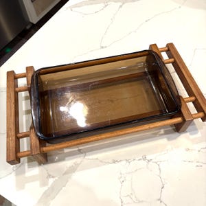 Könnte beinhalten: Ein rechteckiges, bernsteinfarbenes Glas-Backform steht auf einem Serviertablett aus Holz. Das Tablett hat an jedem Ende ein leiterartiges Design mit horizontalen Holzstützen. Die Form ist leer und einsatzbereit.