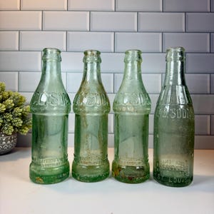 May include: Four vintage green glass soda bottles. The bottles have a classic shape with a narrow neck and a wider base. The bottles have embossed lettering, including the words "C.B. Soda" and "St. Louis". The bottles are displayed on a white surface.