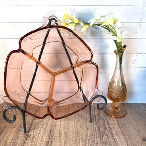 Könnte beinhalten: Ein Vintage-Servierteller aus rosa Glas mit drei Fächern, der von einem schwarzen Metallständer getragen wird. Der Teller hat eine einzigartige, blütenblattartige Form. Rechts befindet sich eine passende rosa Glasvase mit Blumen.