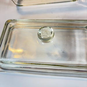 May include: Clear glass rectangular dish with a rounded knob on the lid. The knob has an etched design. The dish is made of clear glass and has a simple, functional design. The dish is likely used for food storage or serving.